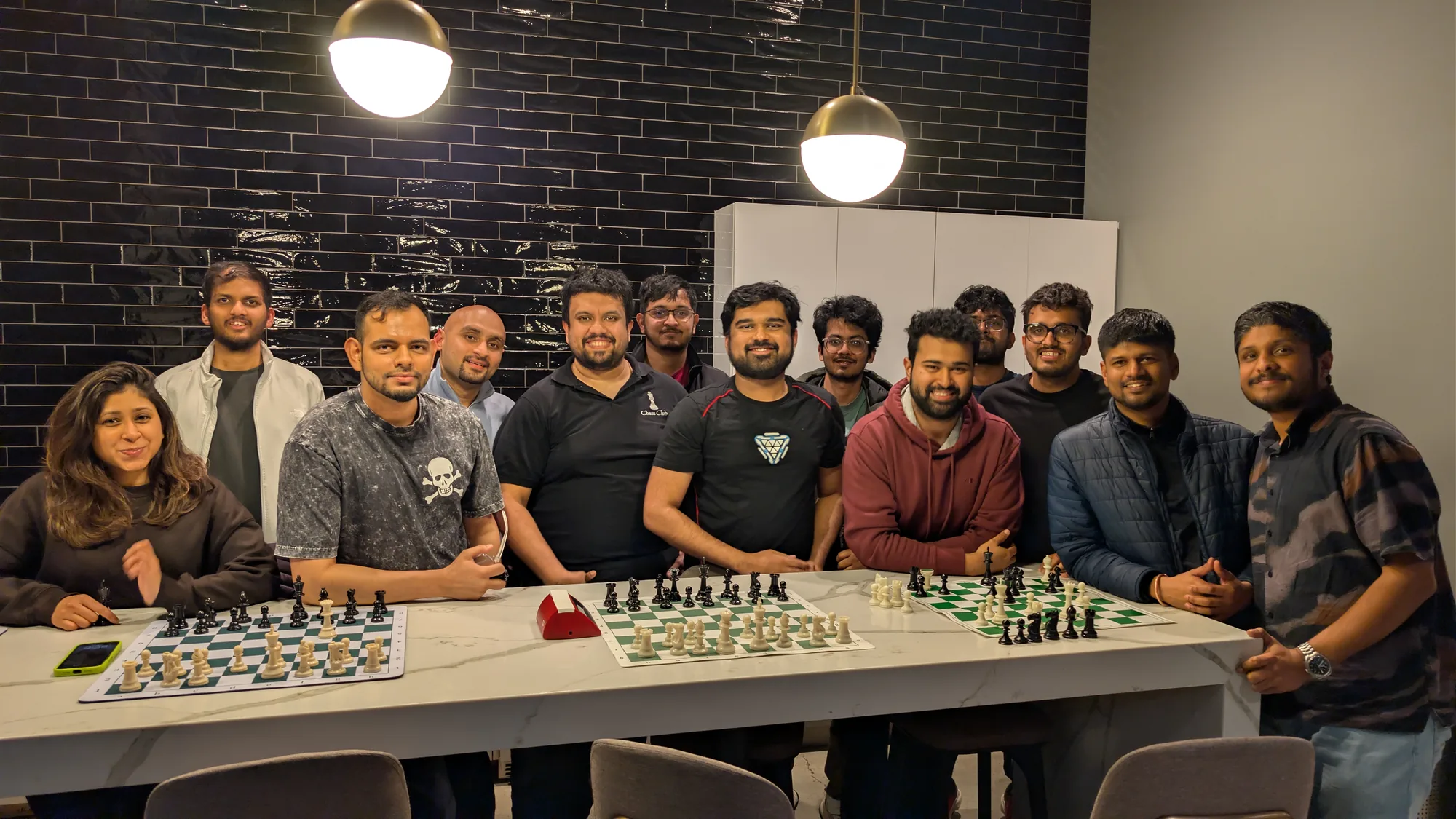 Thirteen club members behind a long marble table with three chess boards and a chess clock, against a black tiled wall.