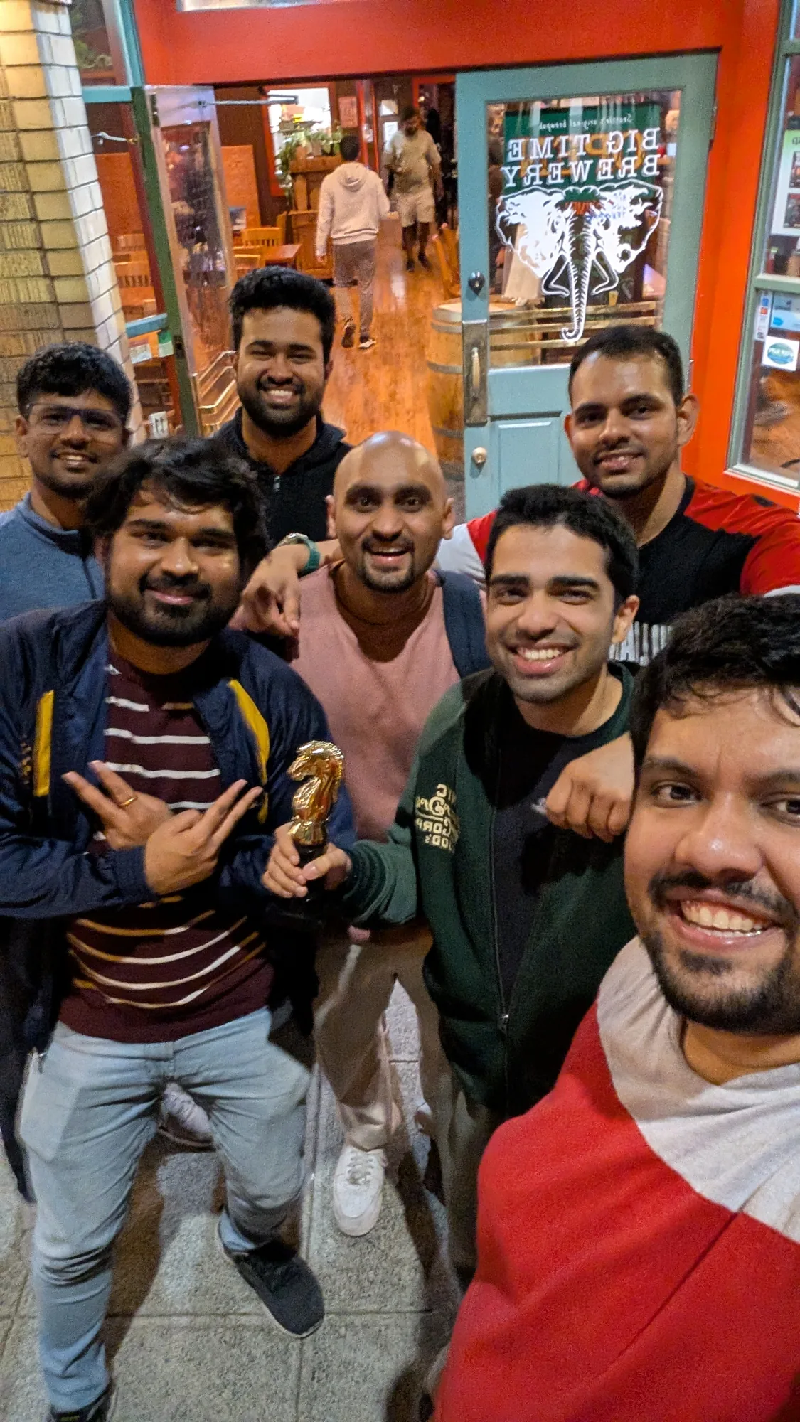Seven club members posing together outside a brewery at night, one holding a small gold knight trophy.